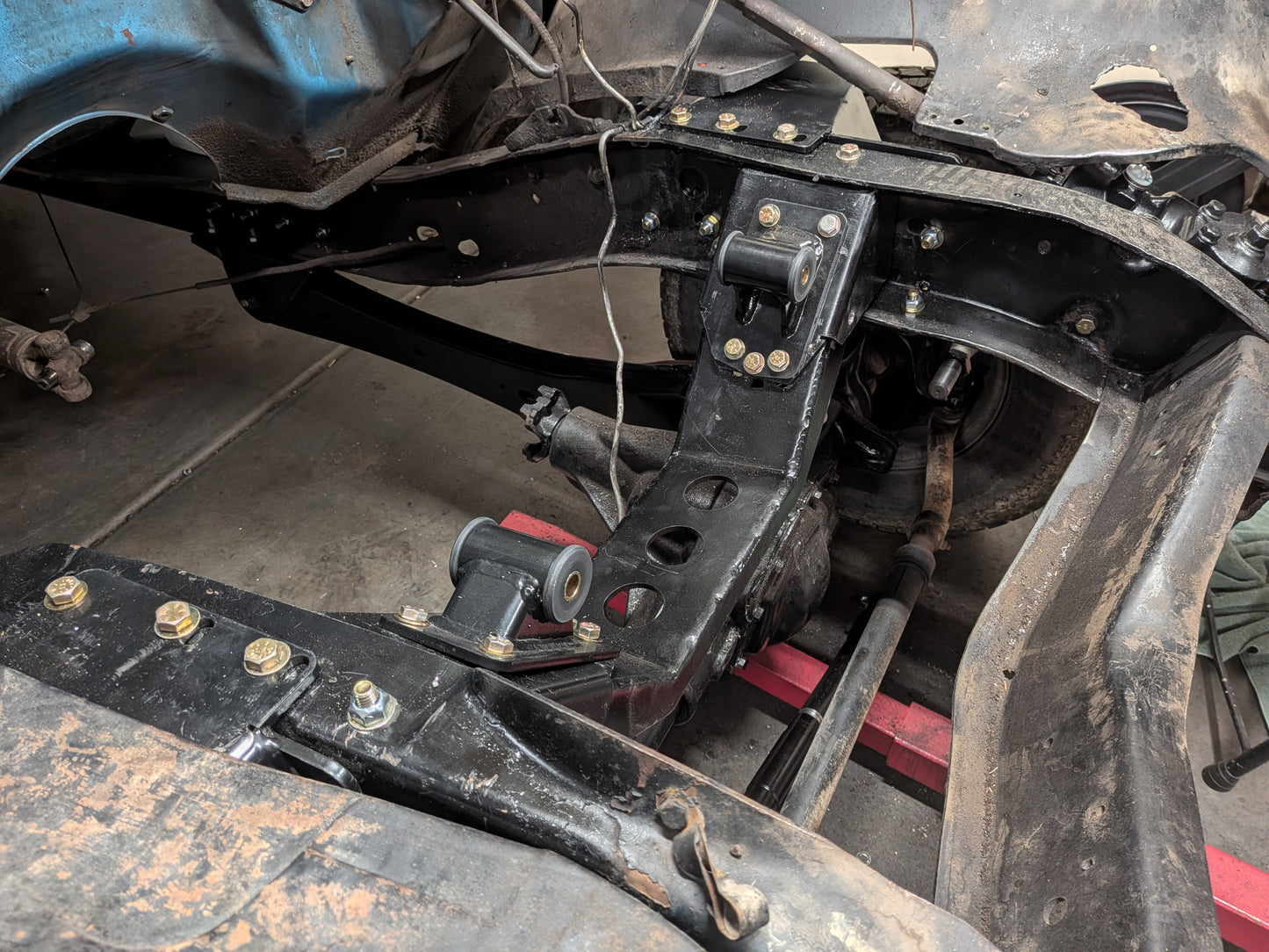 Close-up of a black metal vehicle suspension and chassis component, including a control arm, bushing, and mounting brackets with bolts, photographed in a workshop setting.
