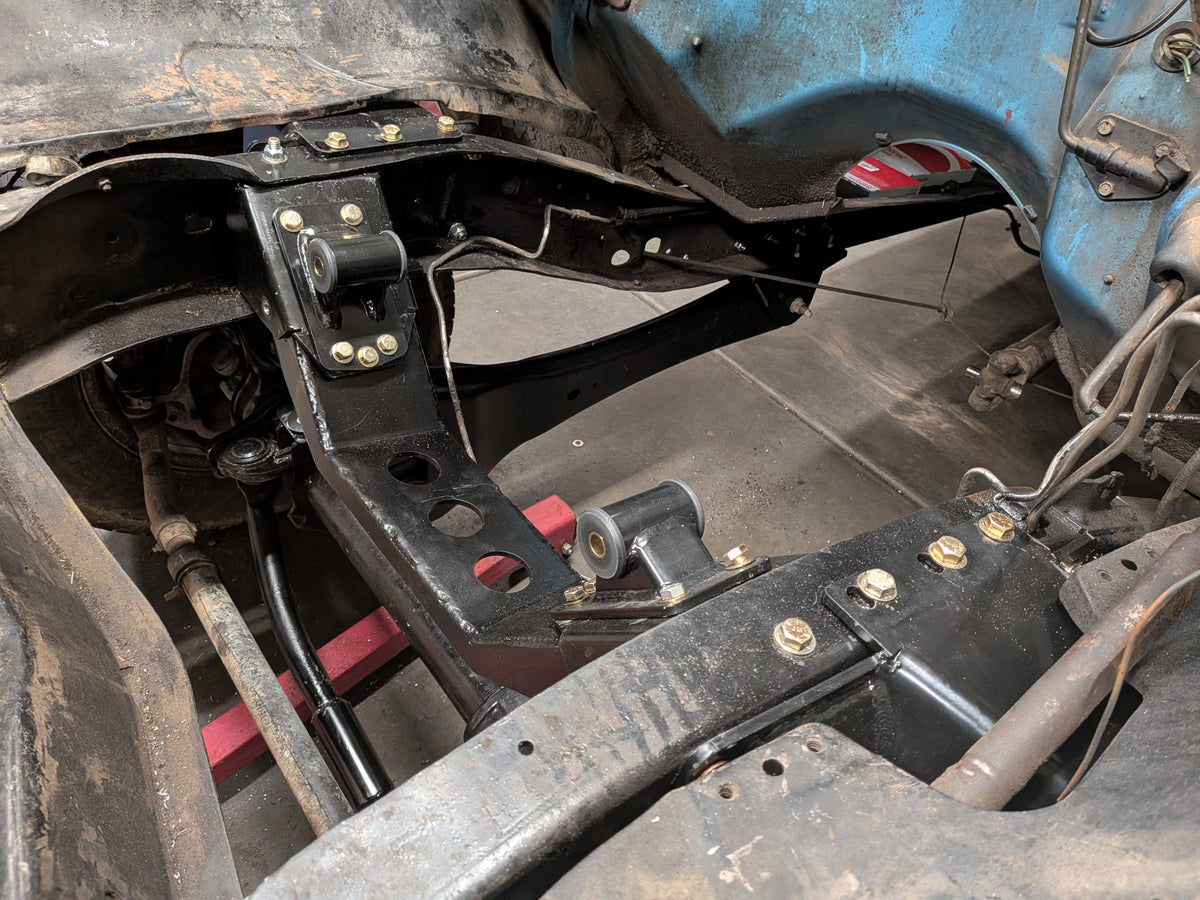 Close-up of a black metal vehicle frame or chassis support beam installed inside a blue car body, with visible bolts, brackets, wiring, and surrounding automotive parts.