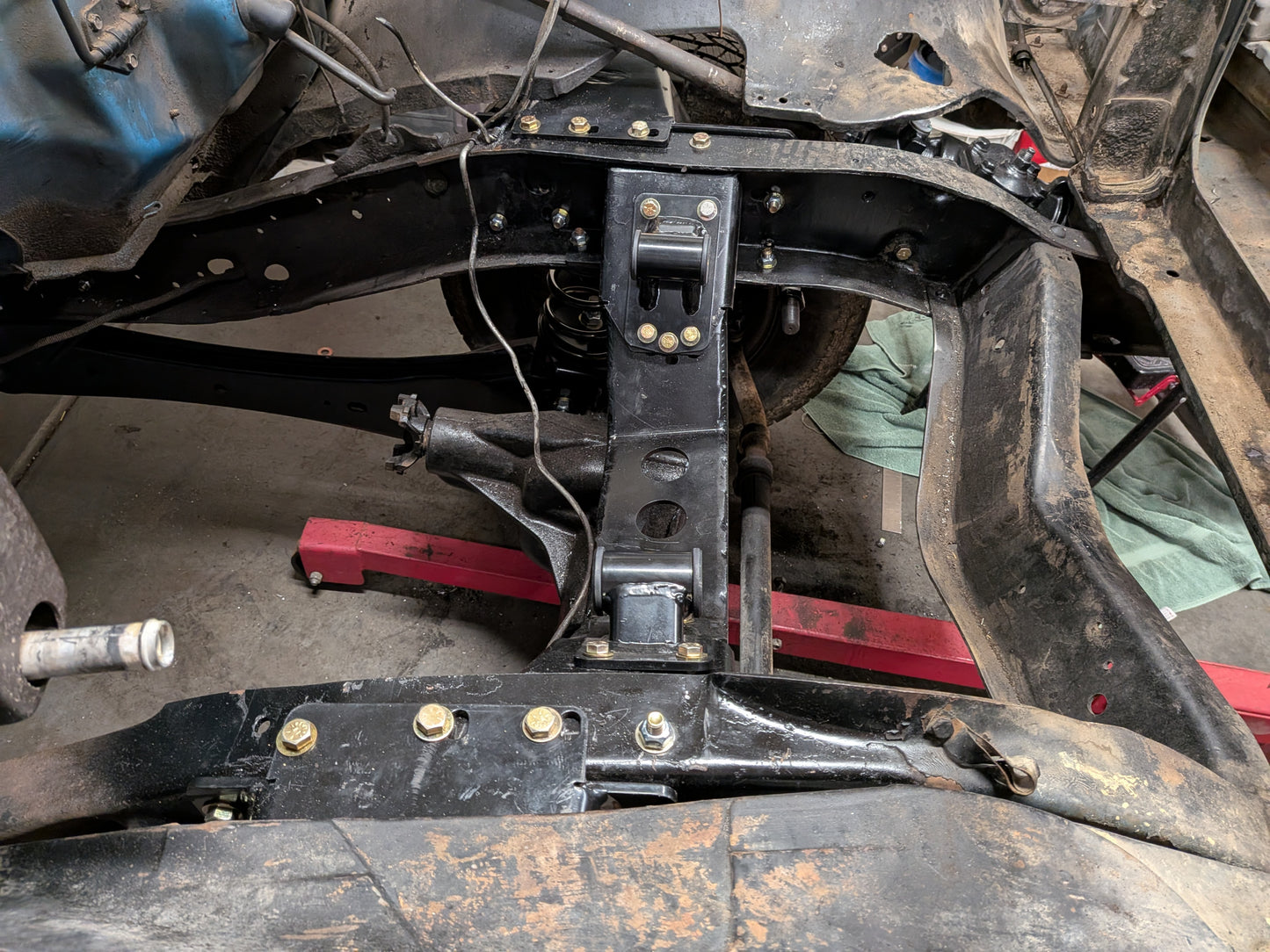 A close-up of a black metal vehicle frame or chassis support beam with visible bolts and mounting points, photographed inside a vehicle bay with other car parts and a red support visible underneath.