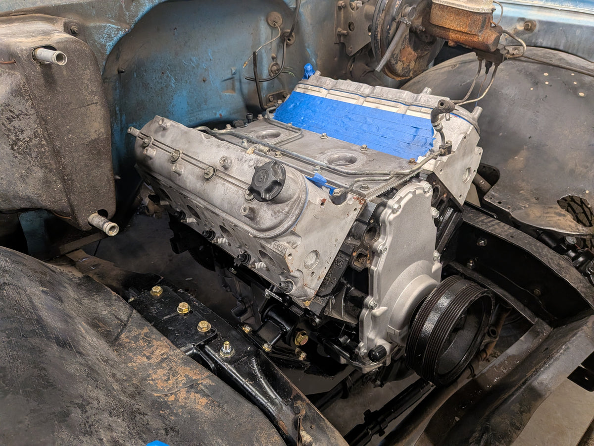 A used silver V8 engine block with a bright blue valve cover is shown installed inside a vehicle frame.