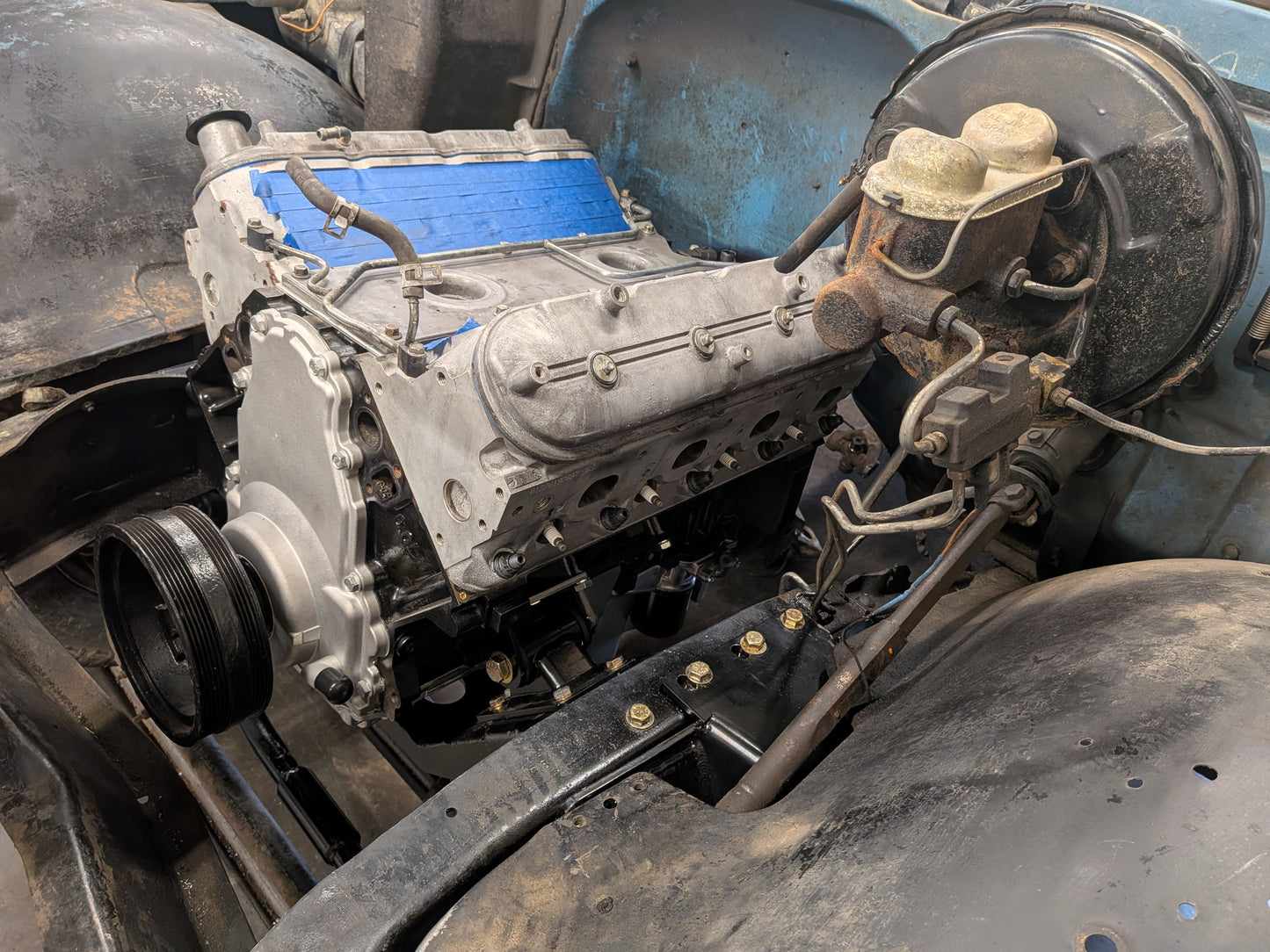 A close-up of a used V8 engine block with a silver aluminum block, a bright blue valve cover, and a black pulley, set within a vintage blue vehicle frame.