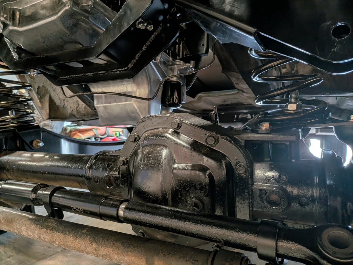 Close-up of a black metal differential housing and surrounding vehicle undercarriage components, including a spring, suspension parts, and pipes, photographed in a workshop setting.